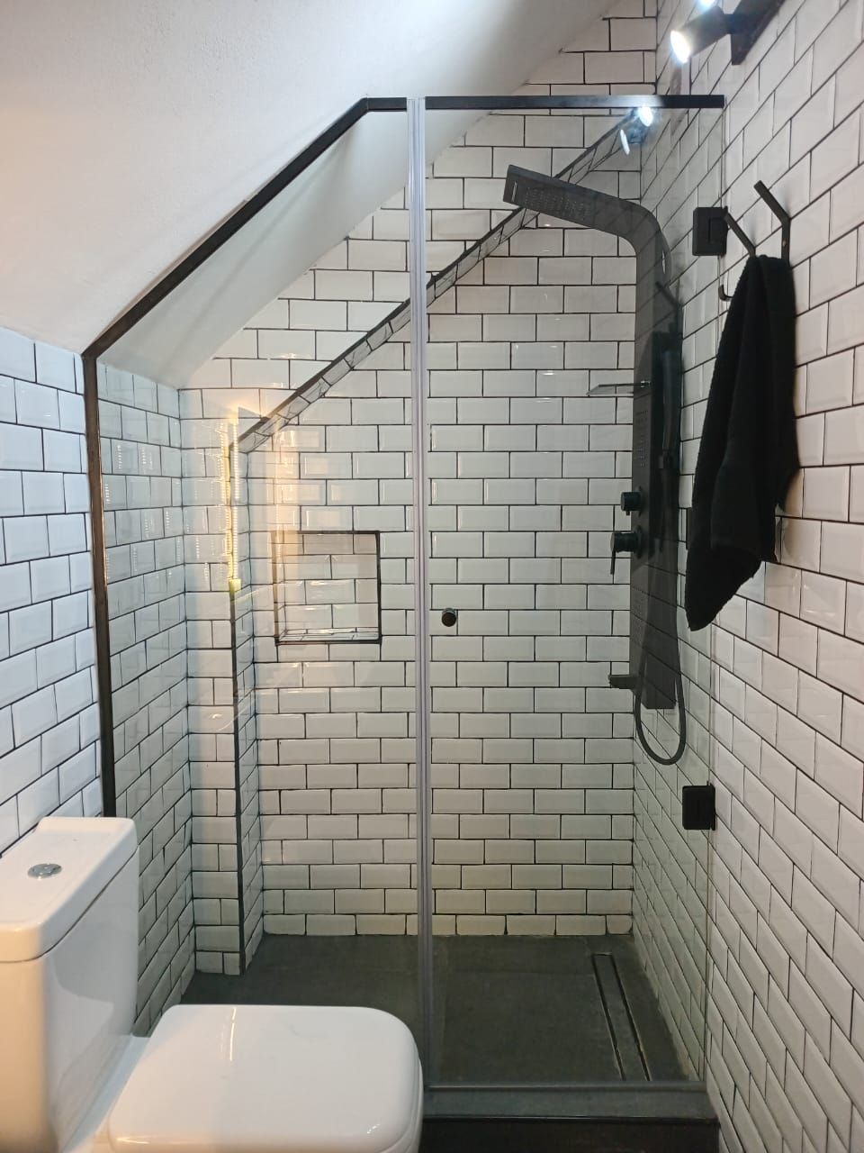 Baño pequeño con azulejos blancos tipo metro, ducha de cristal e inodoro. Grifería de ducha y toallero negros.