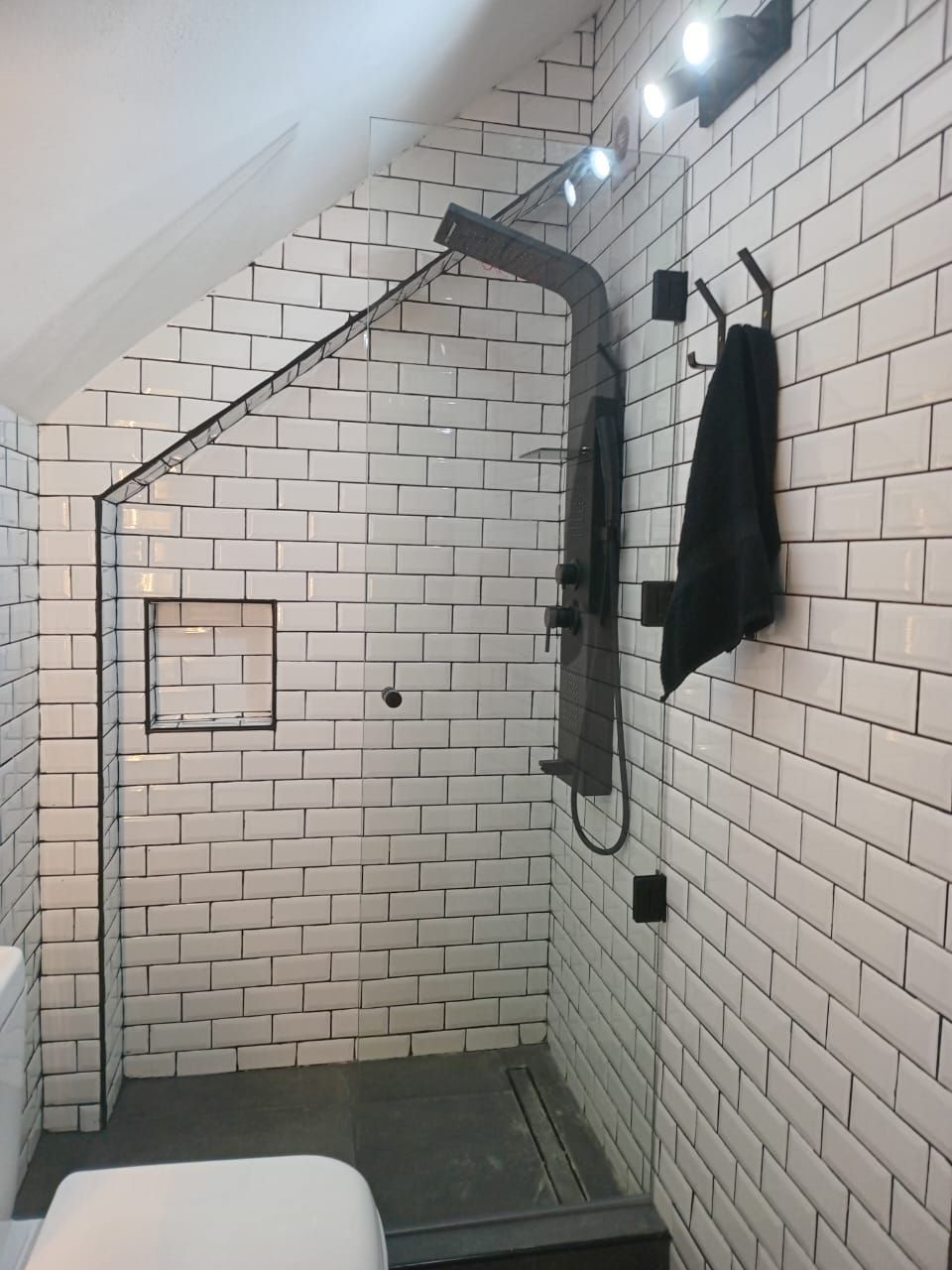 Ducha con azulejos blancos tipo ladrillo, mampara de cristal y grifería negra. Una toalla negra cuelga de la pared.