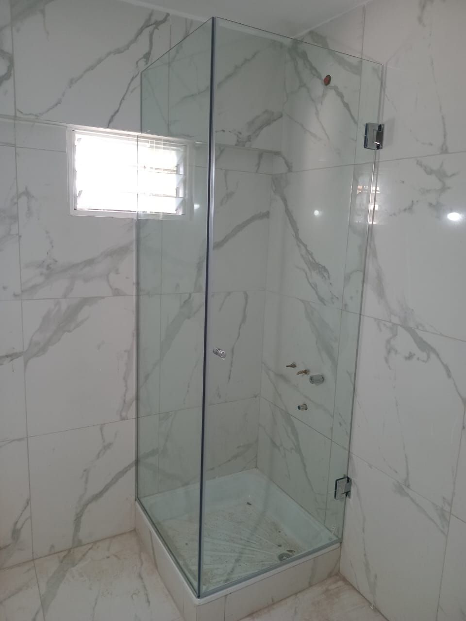 Mampara de ducha de cristal con paredes de azulejos con estampado de mármol blanco y pequeña ventana.