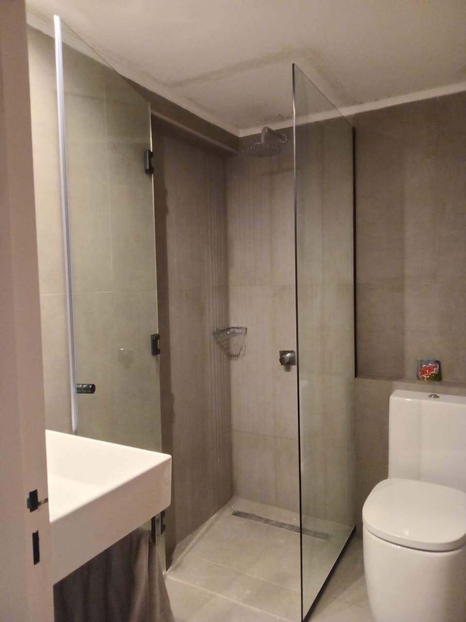 Baño moderno con mampara de ducha de cristal, lavabo y sanitario blancos; paredes y suelo de azulejos grises.