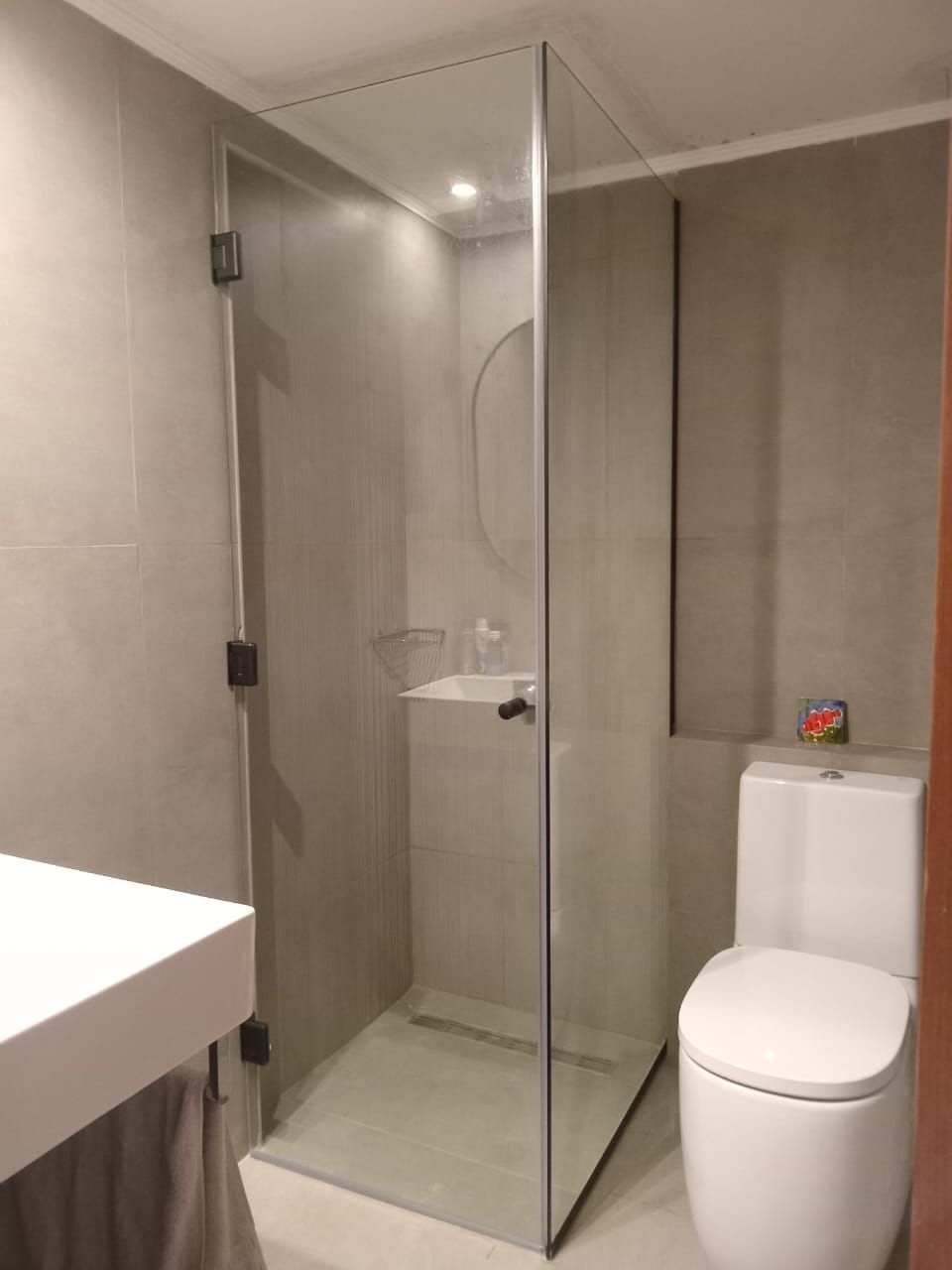 Mampara de ducha de cristal en baño con azulejos grises; inodoro, lavabo y accesorios en color blanco.