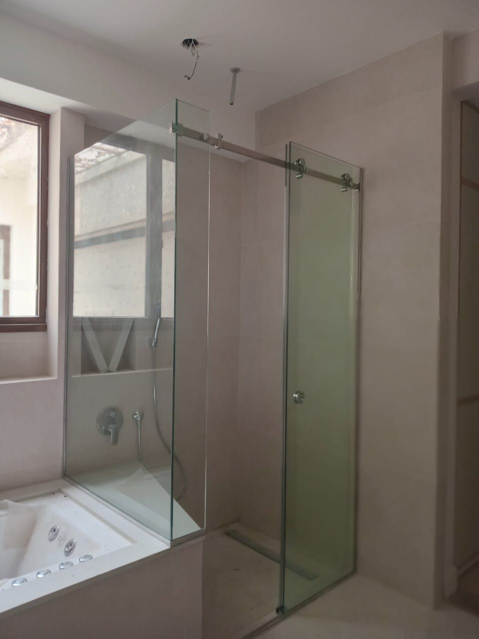 Mampara de ducha con paneles de cristal y puerta corredera junto a una bañera. Paredes beige claro.