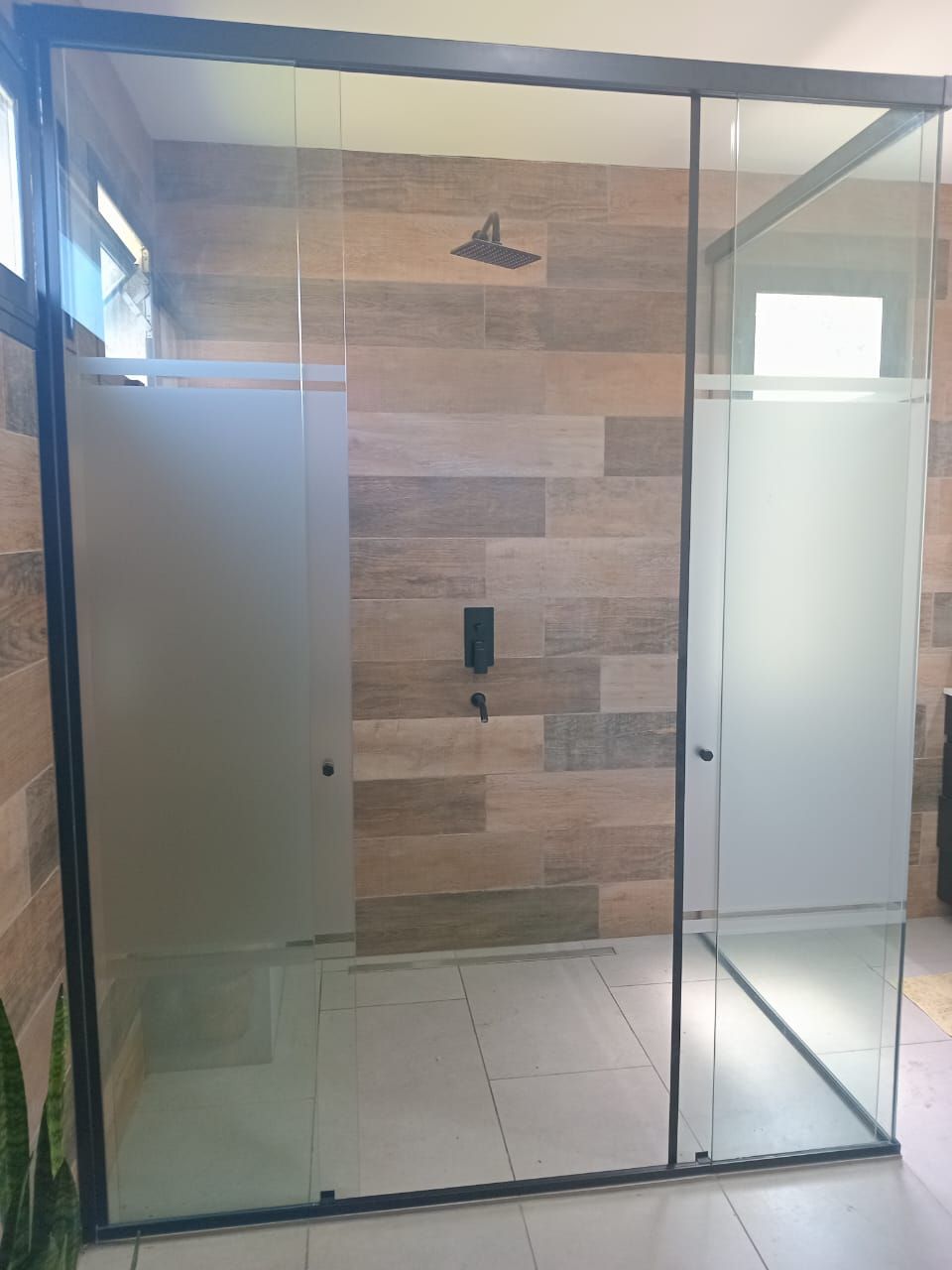 Mampara de ducha de cristal con paneles esmerilados y marco negro sobre una pared de azulejos con aspecto de madera.