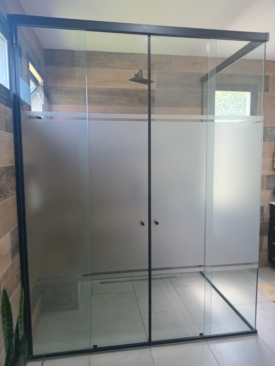 Cabina de ducha de vidrio con marco negro y panel de privacidad esmerilado; sobre azulejos de color marrón claro.