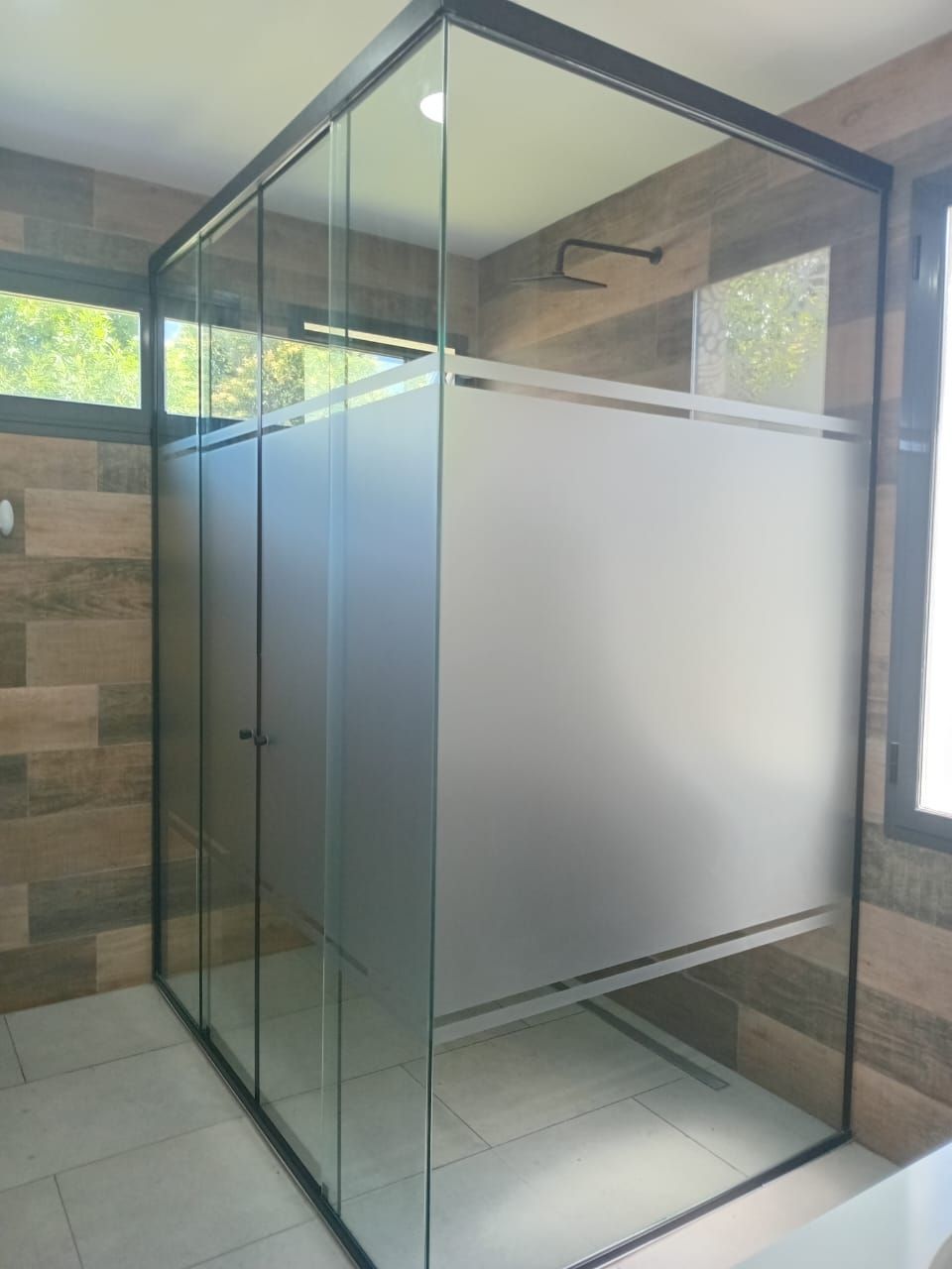 Mampara de ducha de cristal con marco negro y paneles esmerilados sobre paredes revestidas con efecto madera.