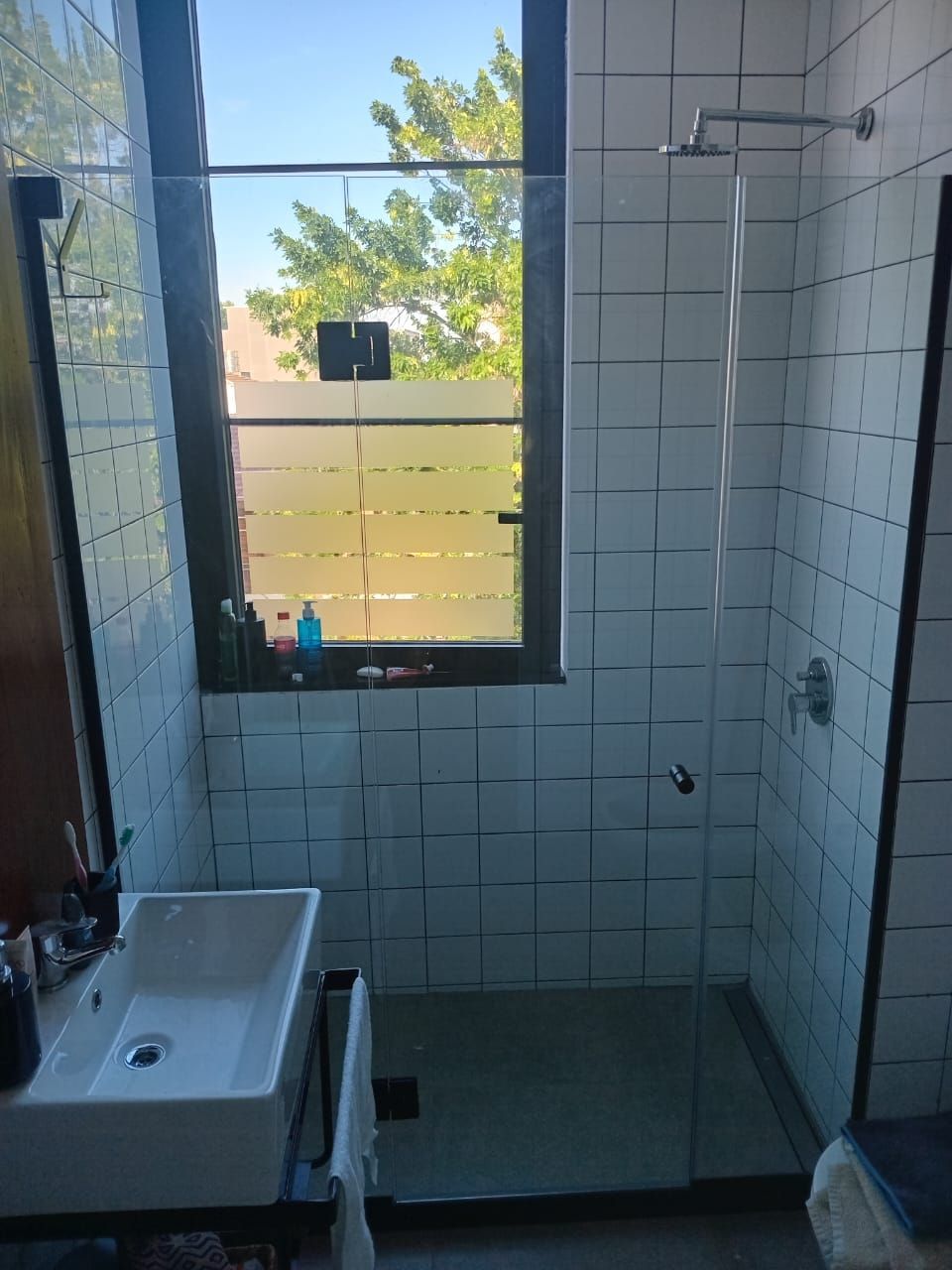 Baño con paredes de azulejos blancos, ducha de cristal, ventana y lavabo.