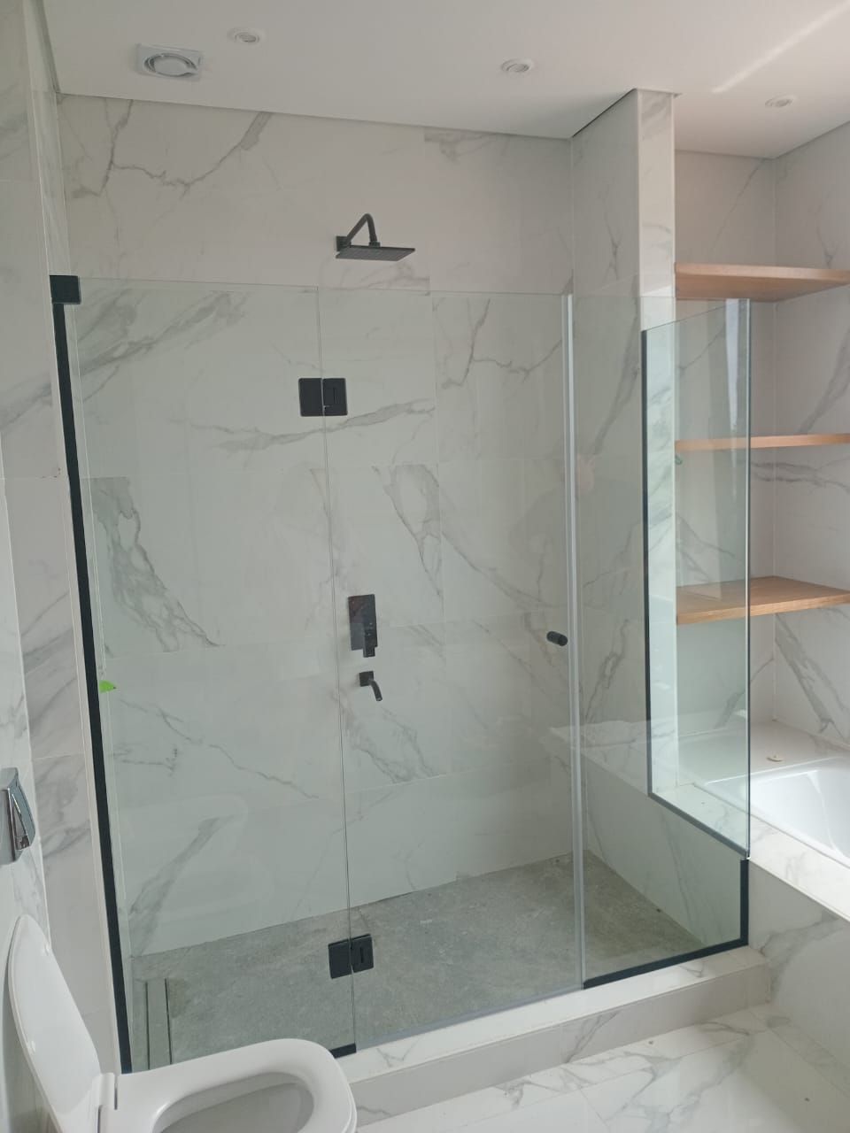 Baño moderno con mampara de ducha de cristal, grifería negra y azulejos imitación mármol. Inodoro blanco en primer plano.