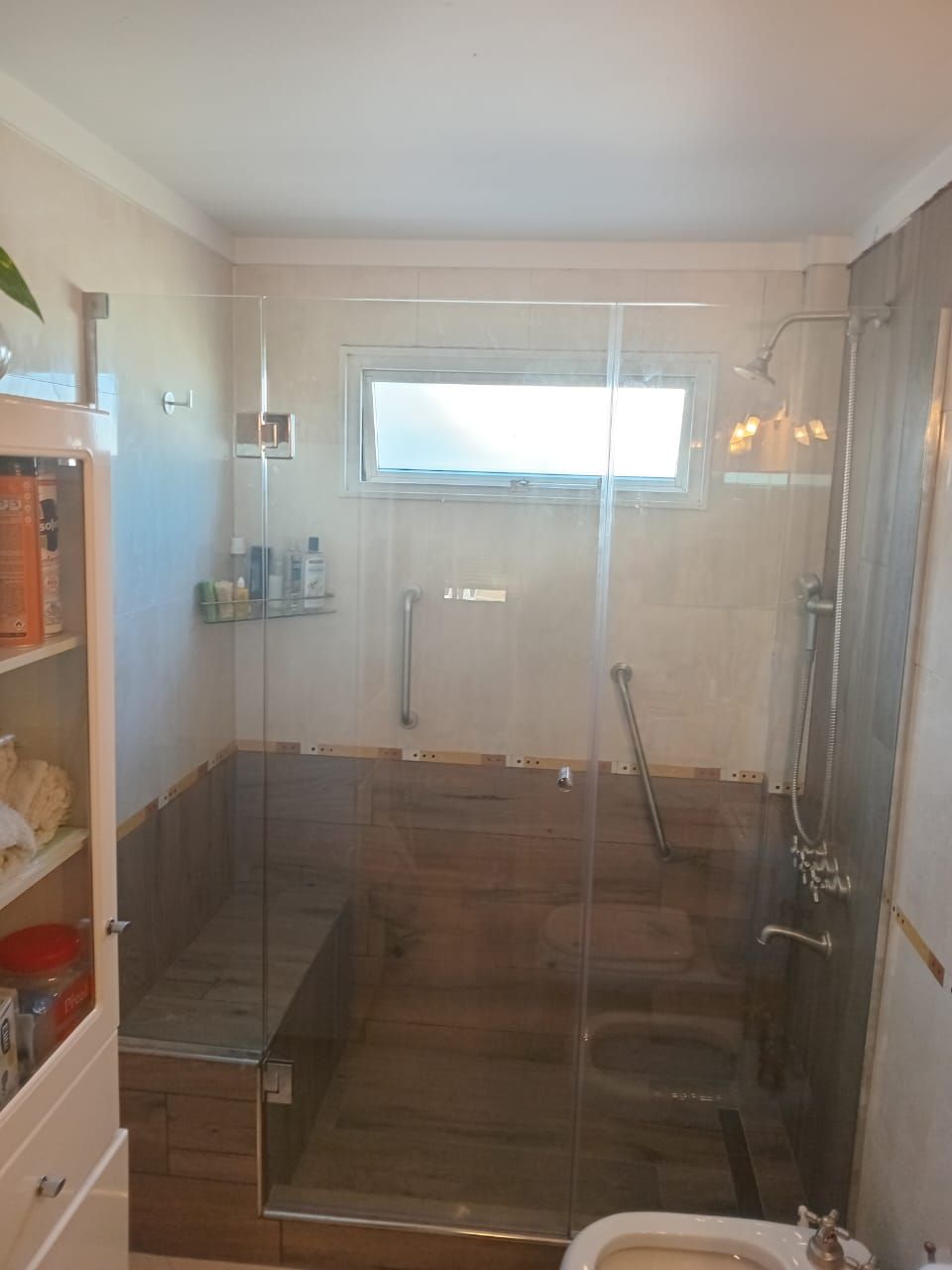 Ducha con puerta de cristal y banco, azulejos marrones, ventana, accesorios cromados.