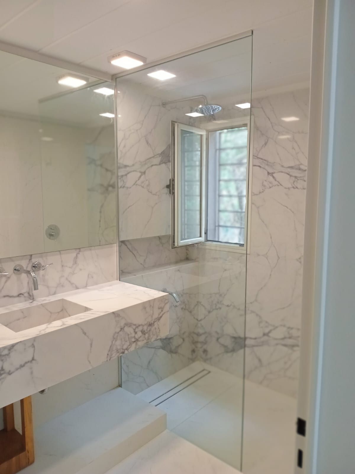 Baño de mármol con mampara de ducha de cristal, ventana y lavabo flotante.