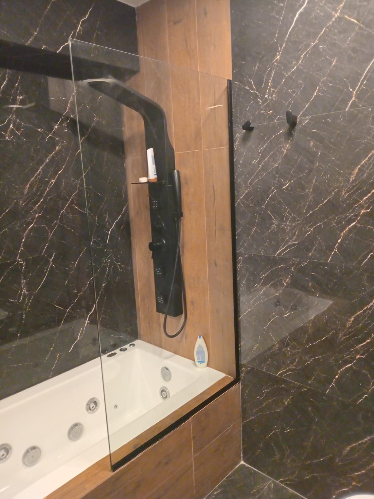 Baño con azulejos de mármol negro, mampara de ducha de cristal y bañera de hidromasaje. Se ve un panel de ducha negro.