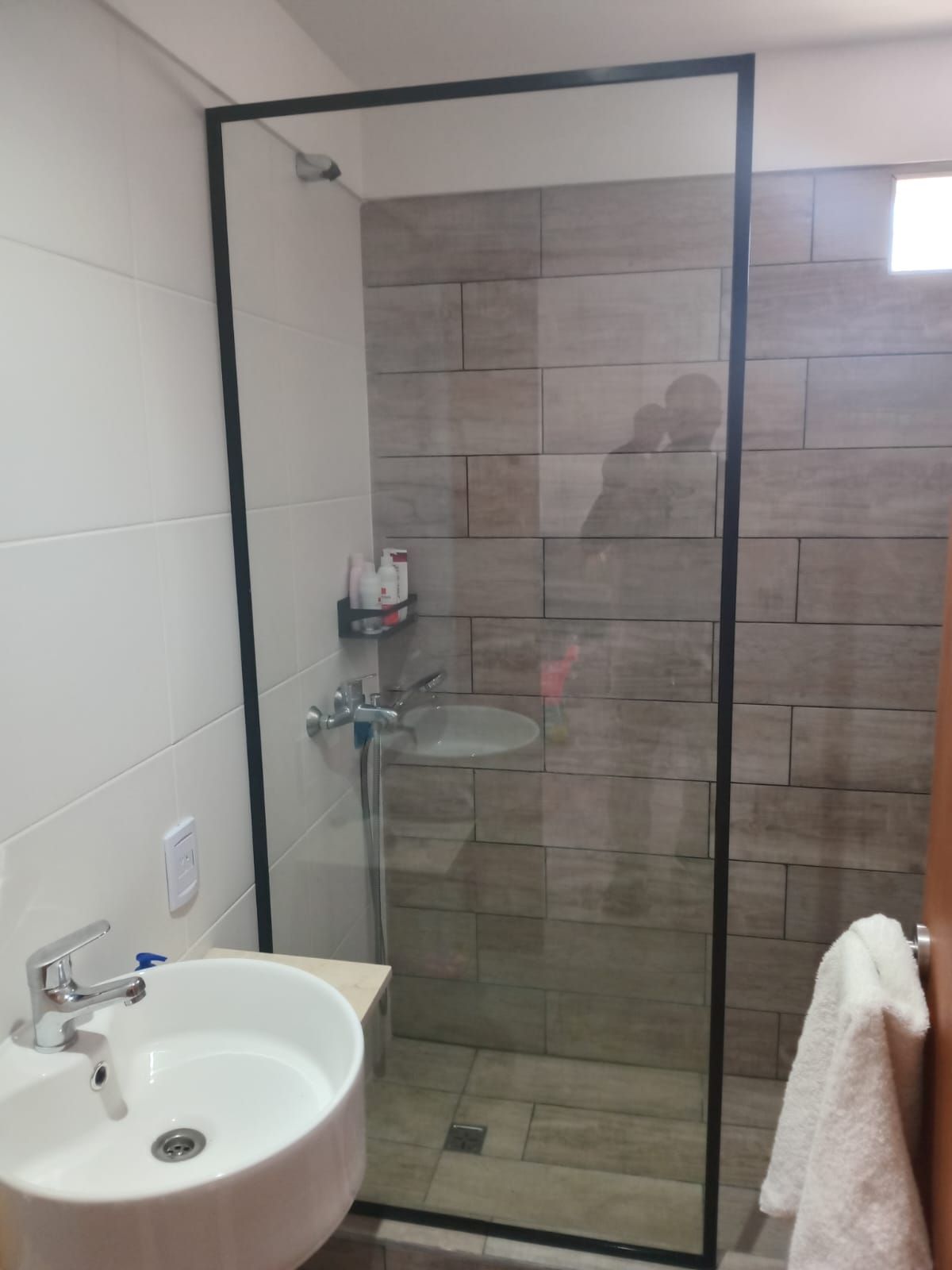 Baño con mampara de ducha de cristal, lavabo blanco y paredes de azulejos de color marrón claro.