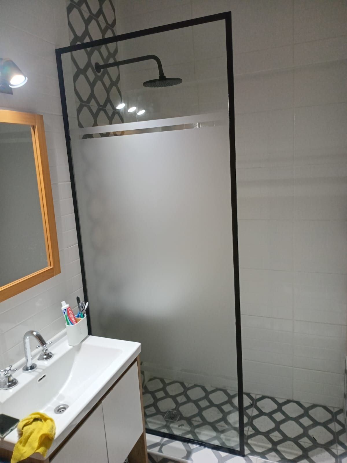 Baño con cabina de ducha, marco negro, vidrio esmerilado, baldosas estampadas, tocador blanco y espejo.
