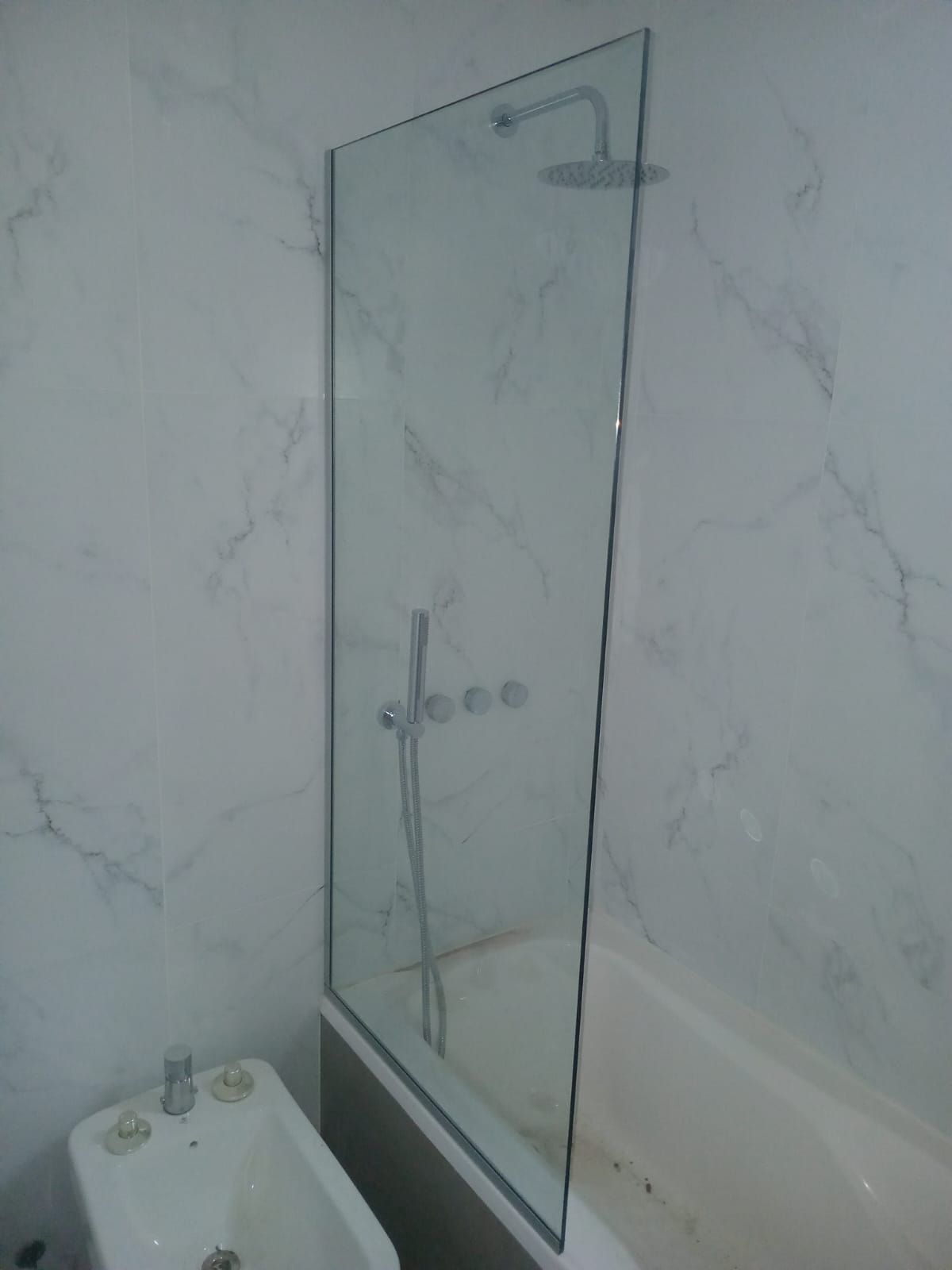 Zona de ducha con mampara de cristal, bañera blanca y paredes de azulejos efecto mármol. Hay un bidé en la esquina.