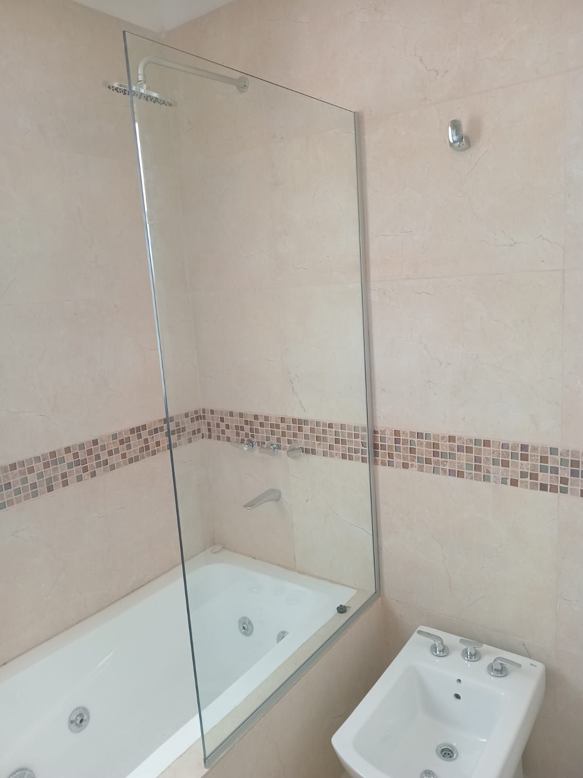 Baño con bañera, mampara de cristal y bidé. Paredes beige con mosaico.