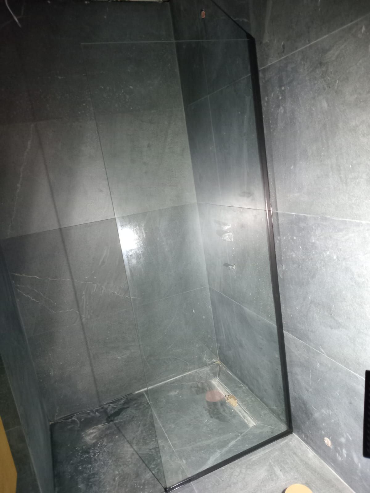 Cabina de ducha con paredes de cristal y suelo de baldosas de color gris oscuro.