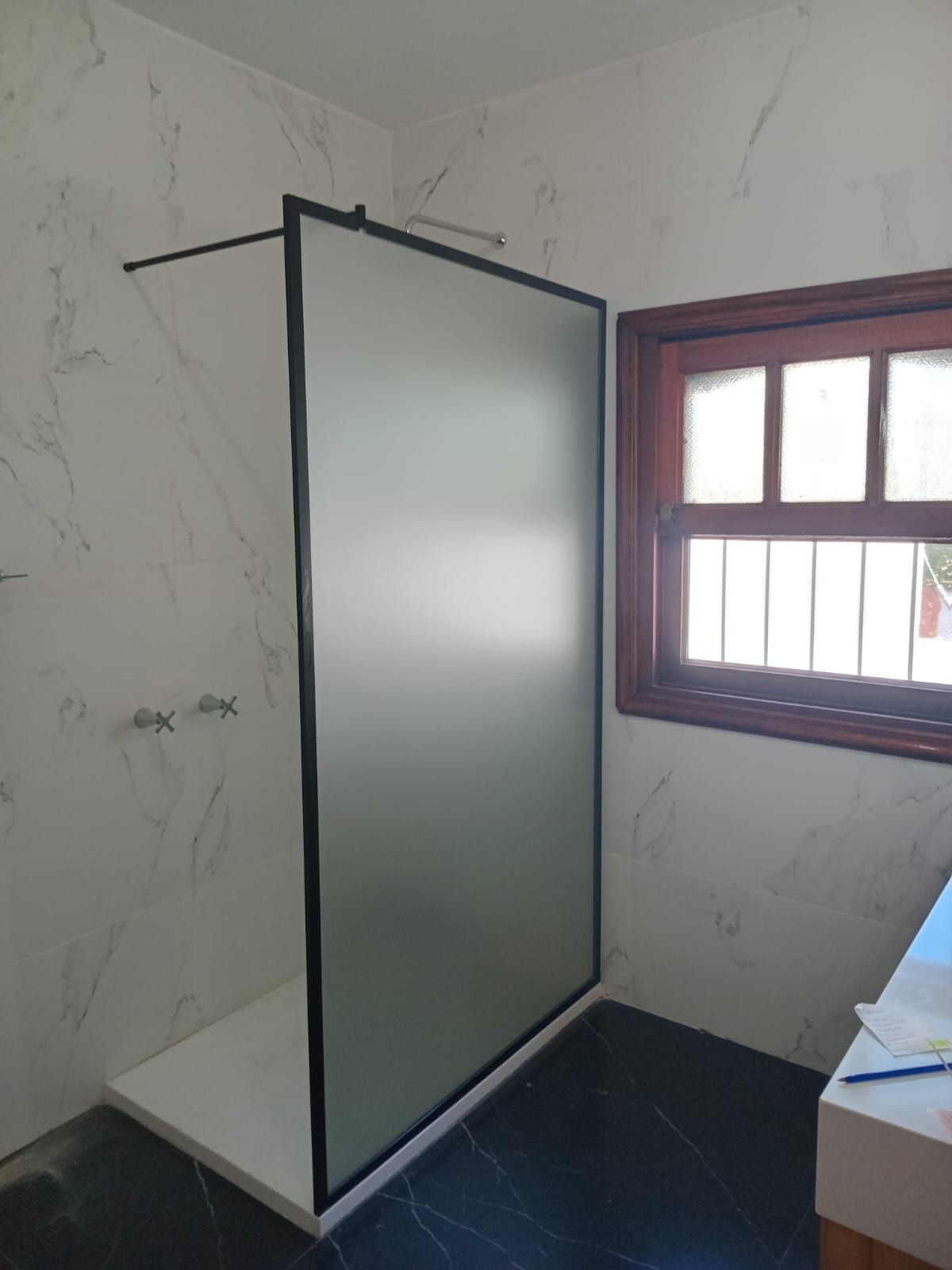 Cabina de ducha con panel de vidrio esmerilado y marco negro junto a una ventana en un baño con azulejos de aspecto mármol.