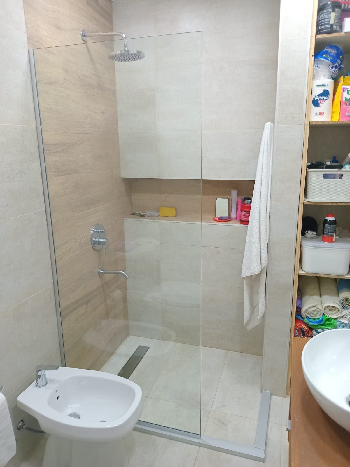 Baño con mampara de ducha de cristal, paredes de azulejos beige, bidé y estantería.