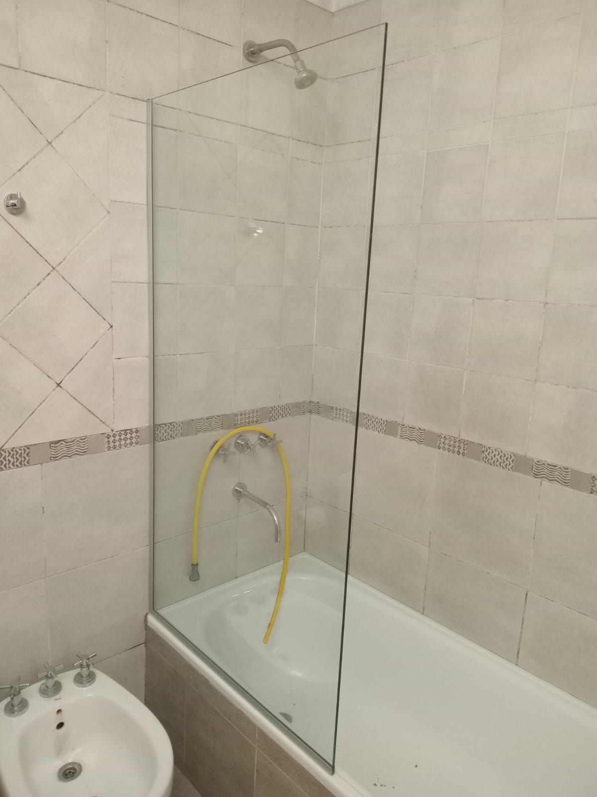 Zona de ducha con mampara de cristal, bañera, rociador y paredes alicatadas.