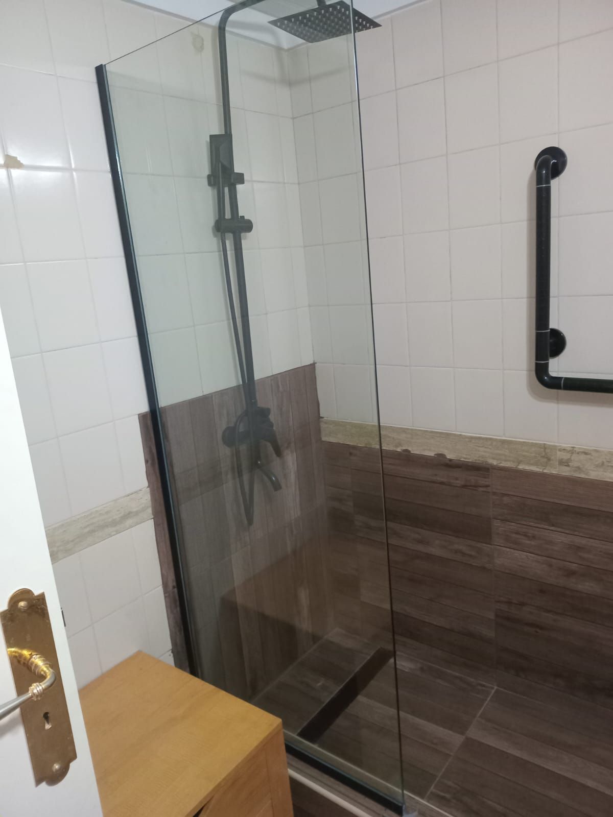 Cabina de ducha con puerta de cristal, accesorios oscuros, azulejos marrones.