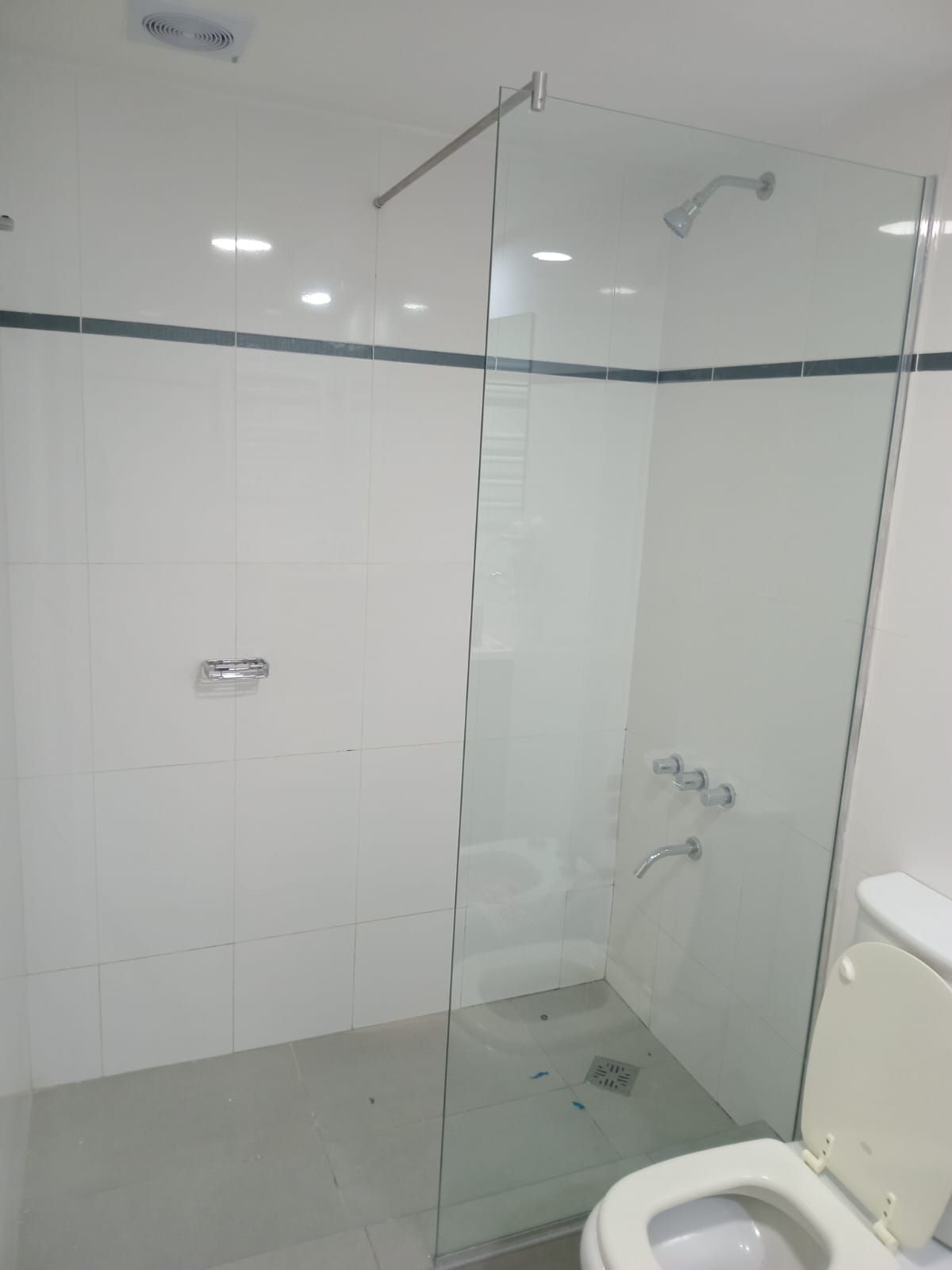 Ducha con azulejos blancos y mampara de cristal. Inodoro en primer plano. Grifería cromada.