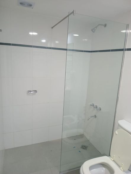 Ducha con azulejos blancos y mampara de cristal. Inodoro en primer plano. Grifería cromada.