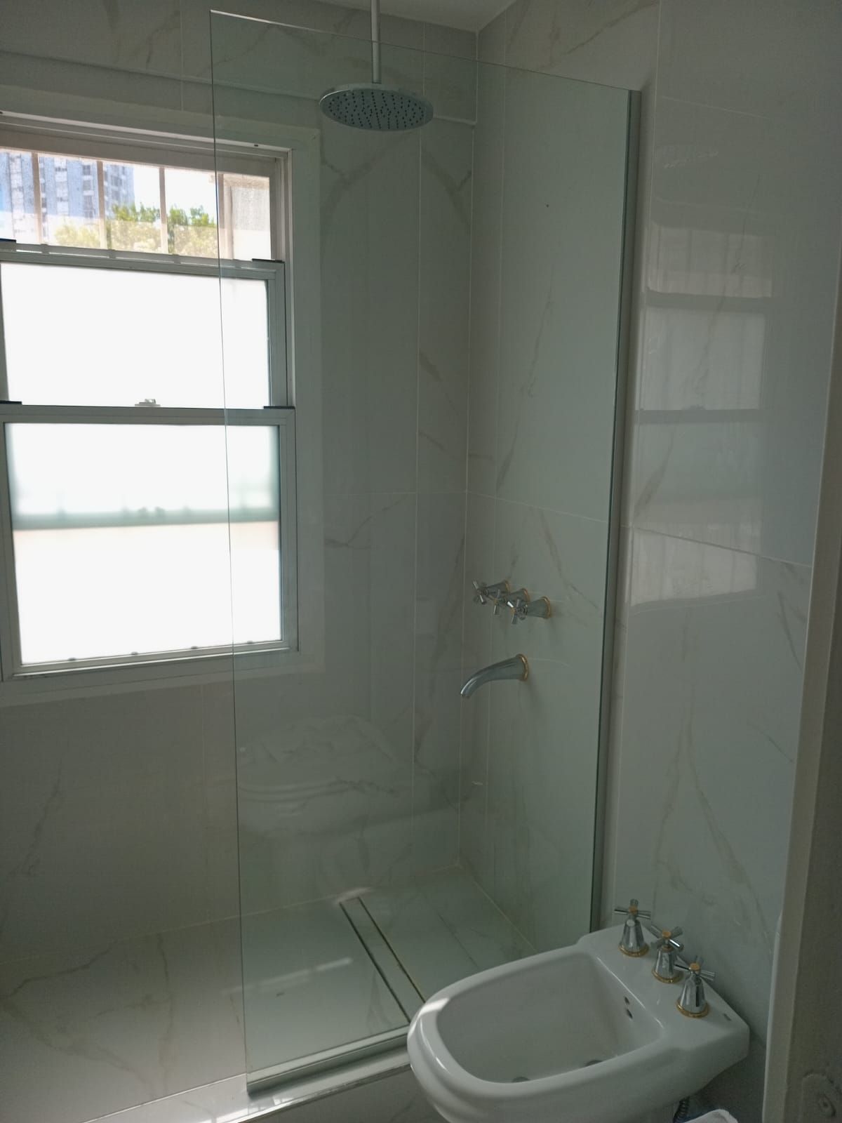 Baño con ducha, mampara de cristal, bidé y ventana. Azulejos y grifería blancos.