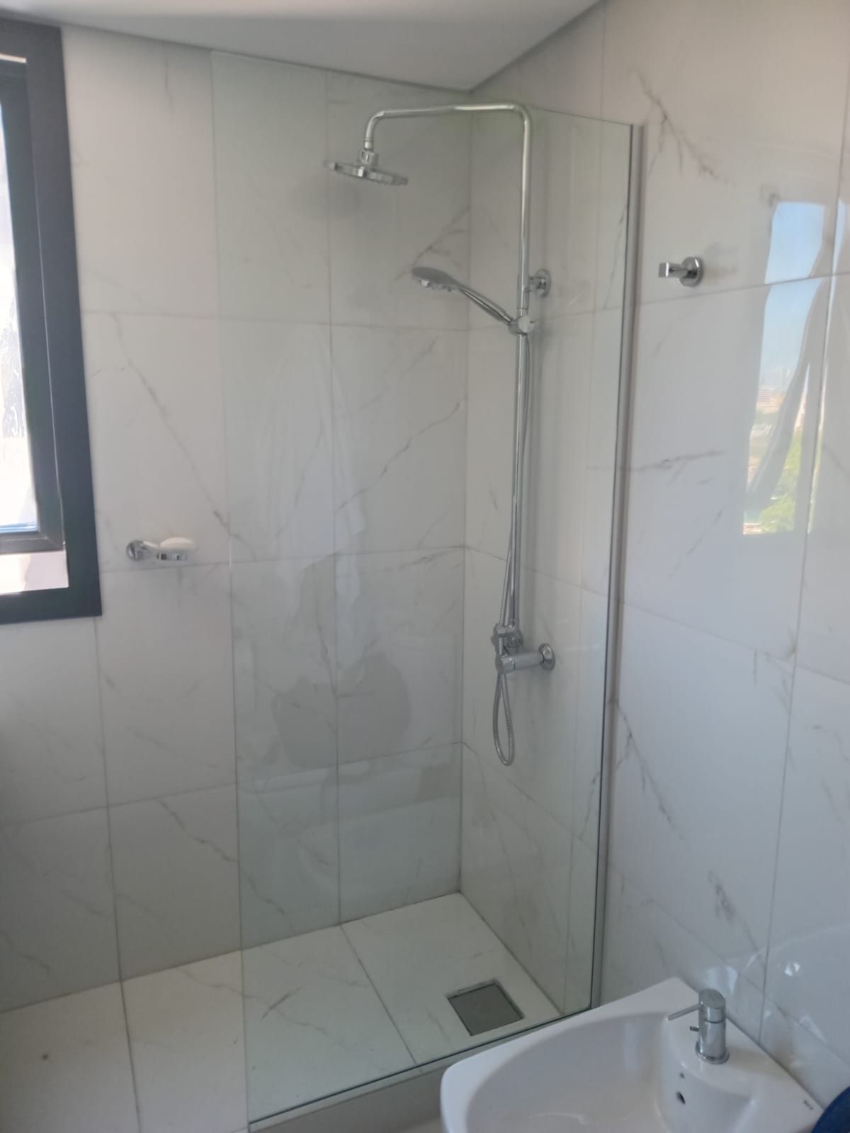 Cabina de ducha con puerta de cristal, baldosas de mármol blanco y accesorios cromados.