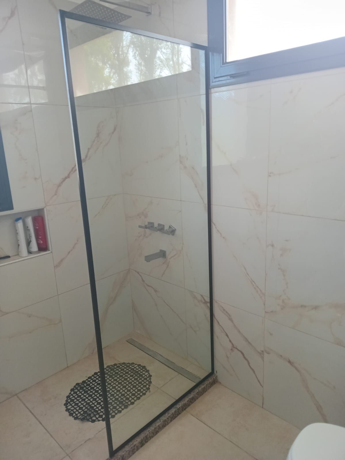 Ducha con puerta de cristal, paredes de azulejos con diseño de mármol y una alfombra de baño ovalada negra en el piso.