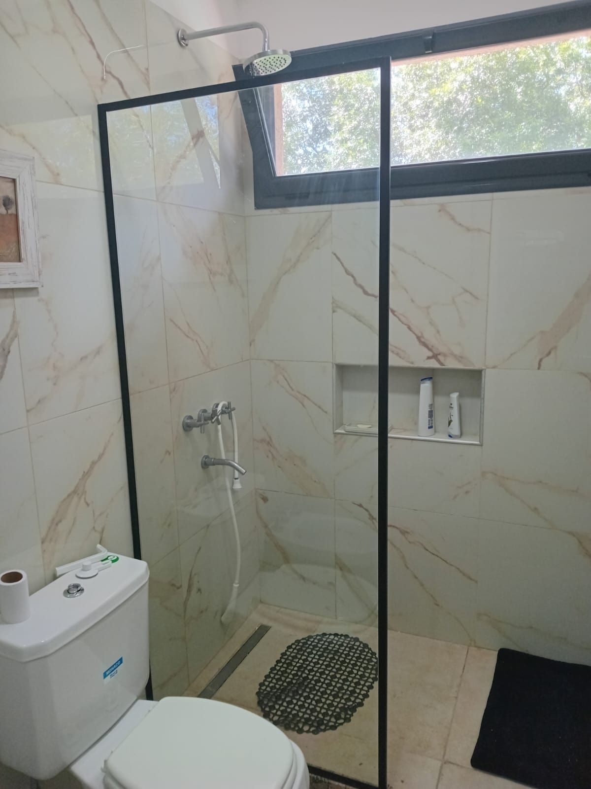 Baño con mampara de ducha de cristal e inodoro. Azulejos de mármol beige y molduras negras.