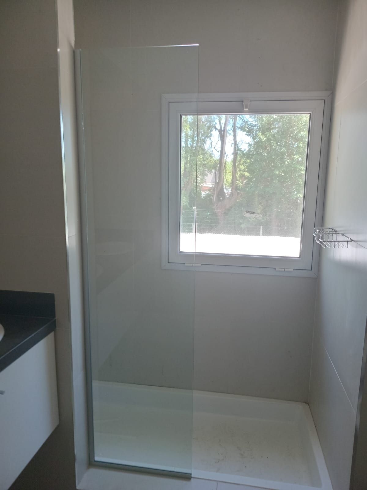 Cabina de ducha con puerta de cristal, paredes blancas y ventana.