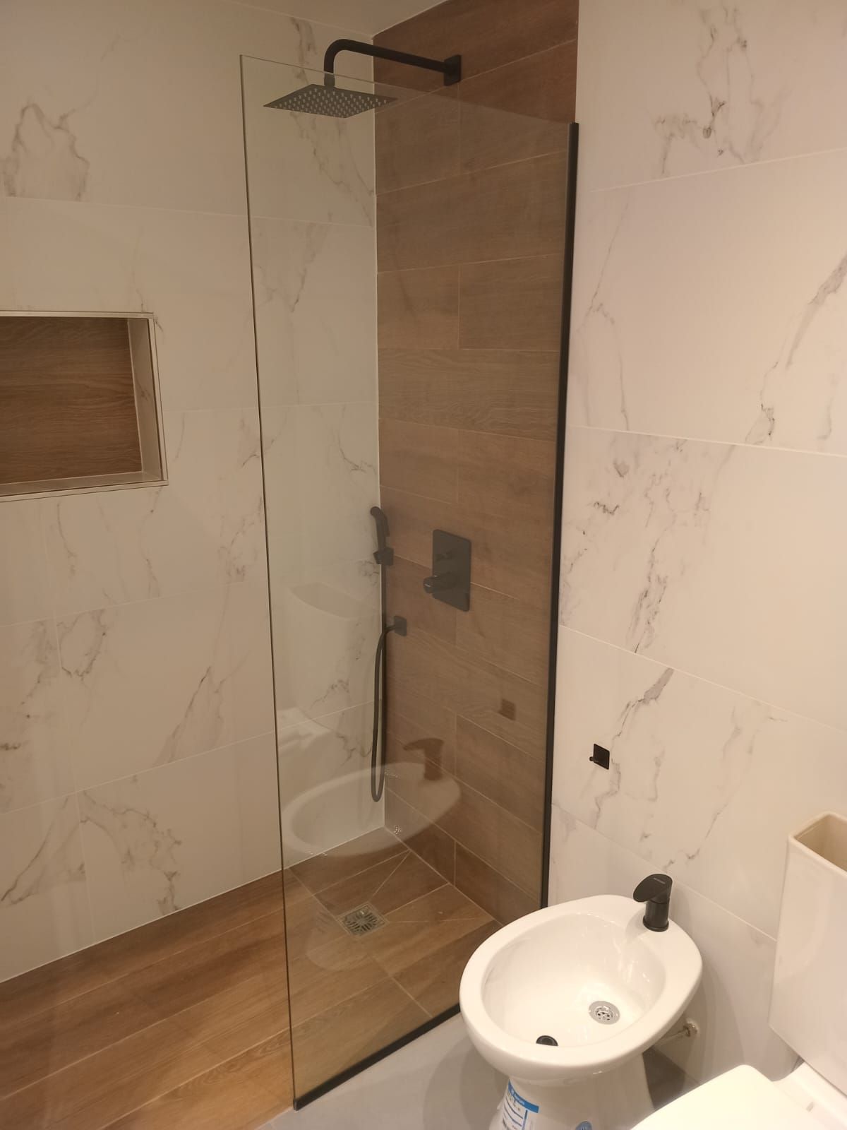 Baño con mampara de ducha de cristal, azulejos imitación madera y paredes de mármol. Se ven grifería negra y un bidé.