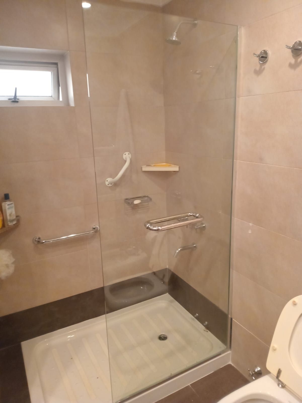 Cabina de ducha accesible con barras de apoyo y jaboneras integradas. Paredes de azulejos beige