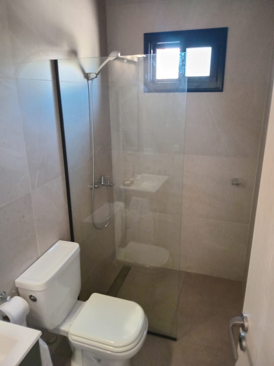 Baño con inodoro, ducha de cristal y ventana pequeña. Paredes de azulejos beige.