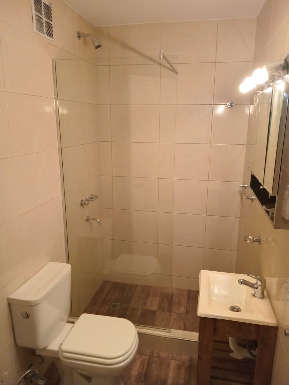 Baño con ducha de cristal, inodoro, lavabo y suelo imitación madera. Paredes beige y espejo.