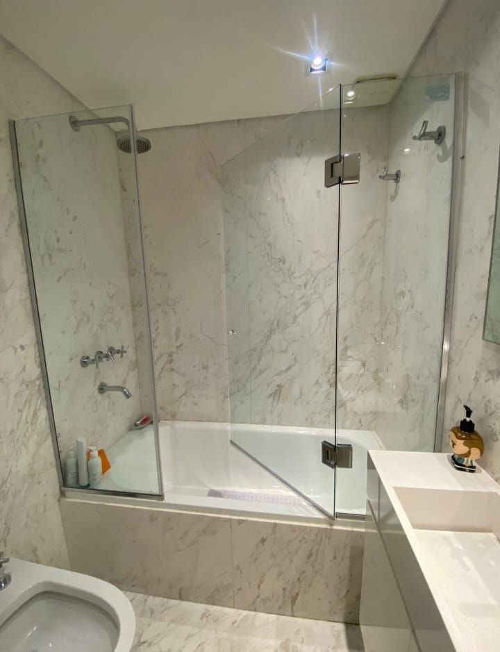 Baño con paredes de mármol y ducha con mampara de cristal sobre la bañera. El lavabo y el inodoro blancos