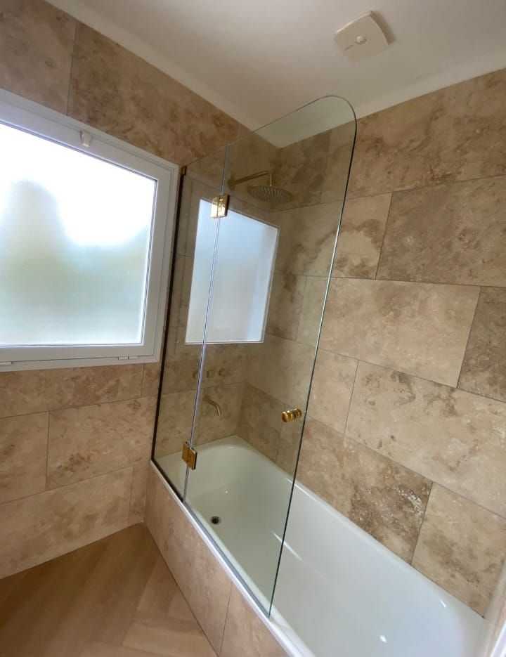 Baño con mampara de cristal, bañera blanca, azulejos beige y ventana esmerilada.