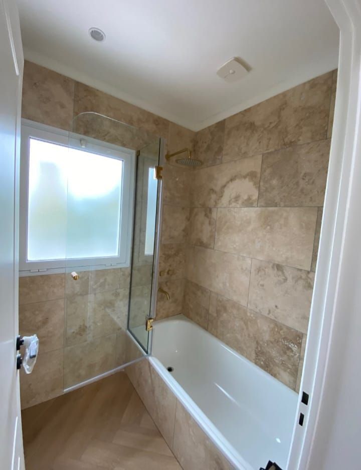 Baño con paredes de azulejos beige, bañera blanca y mampara de cristal. Ventana esmerilada e iluminación empotrada.