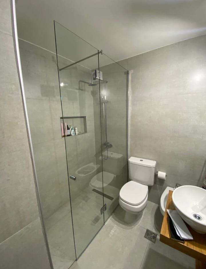 Baño moderno con mampara de ducha de cristal, inodoro, lavabo y azulejos de color gris neutro.