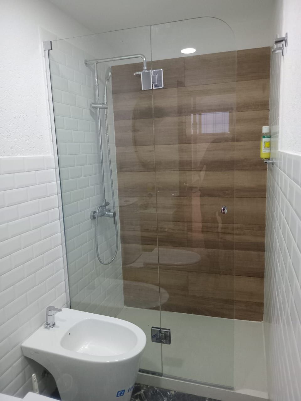 Cabina de ducha con mampara de cristal, bidé y pared revestida de madera. Paredes de azulejos blancos 