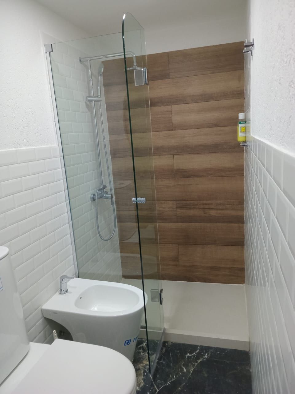 Baño con puerta de ducha de cristal, pared imitación madera, azulejos blancos, inodoro y bidet.