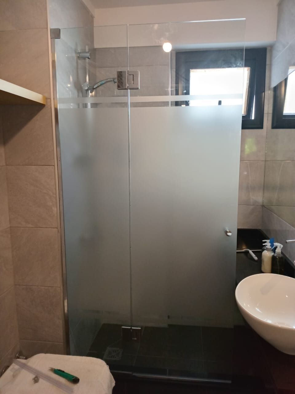 Baño con mampara de ducha de cristal, lavabo y ventana. La mampara de la ducha está parcialmente esmerilada.