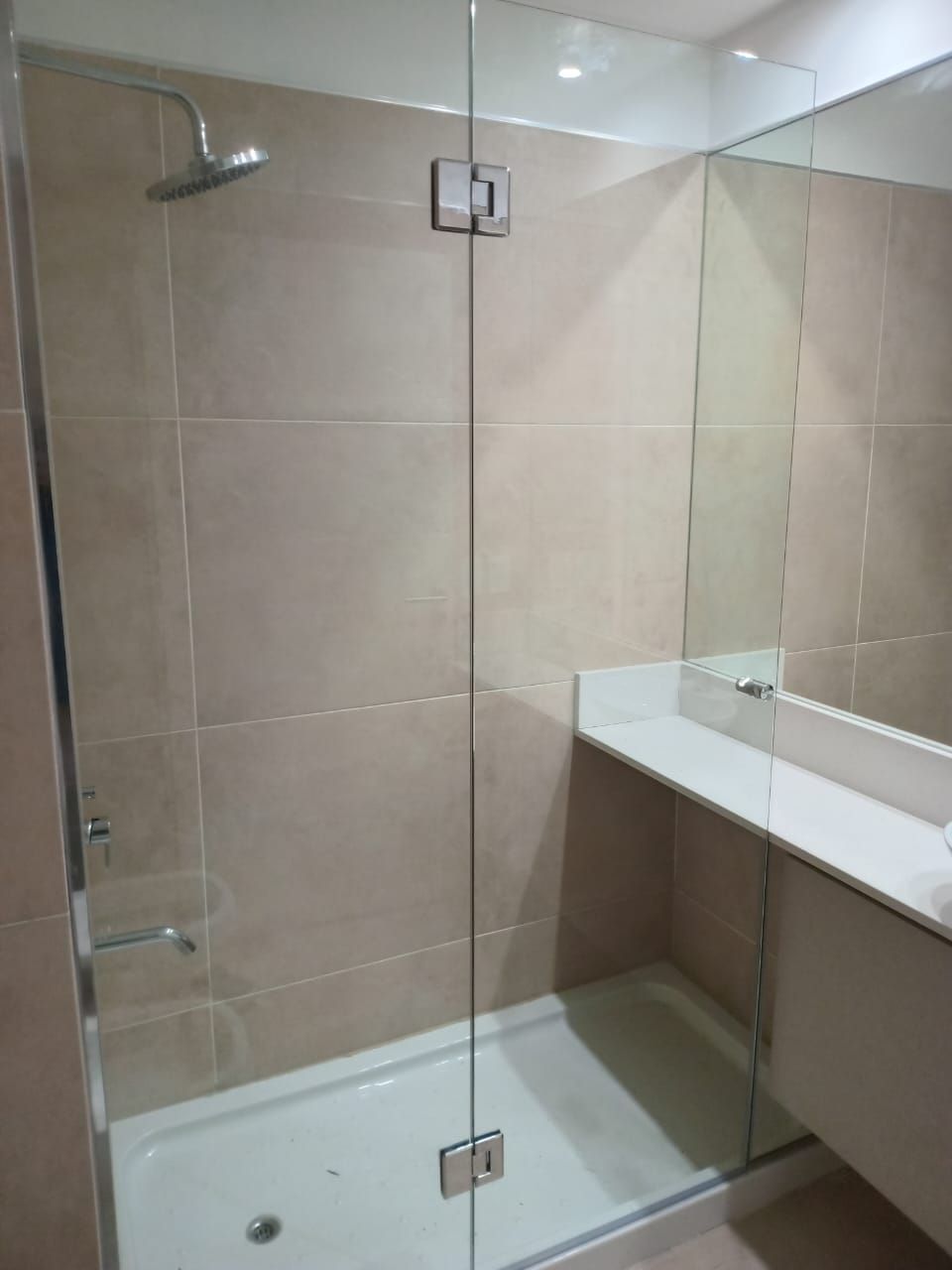 Mampara de ducha de cristal con paredes revestidas de azulejos beige, base de ducha blanca y tocador.