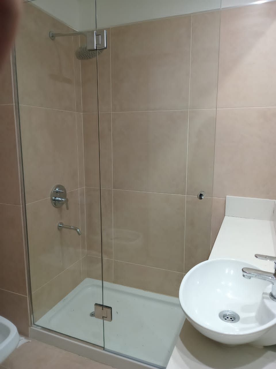 Baño con mampara de ducha de cristal, azulejos beige, lavabo blanco sobre encimera.