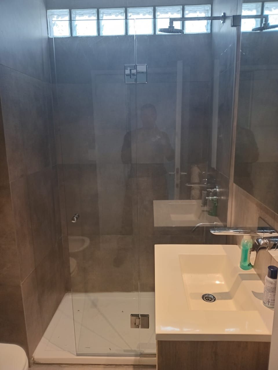 Baño con ducha, lavabo y ventana de bloques de vidrio. Paredes de azulejos marrones y grifería blanca.