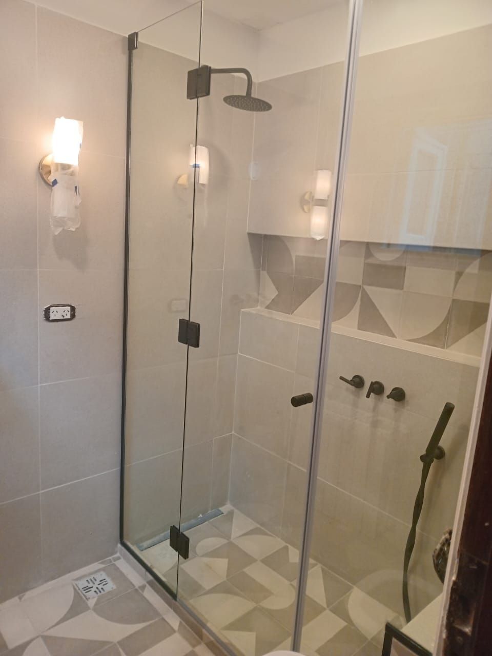 Ducha de cristal con grifería negra y suelo estampado. Paredes gris claro con apliques.