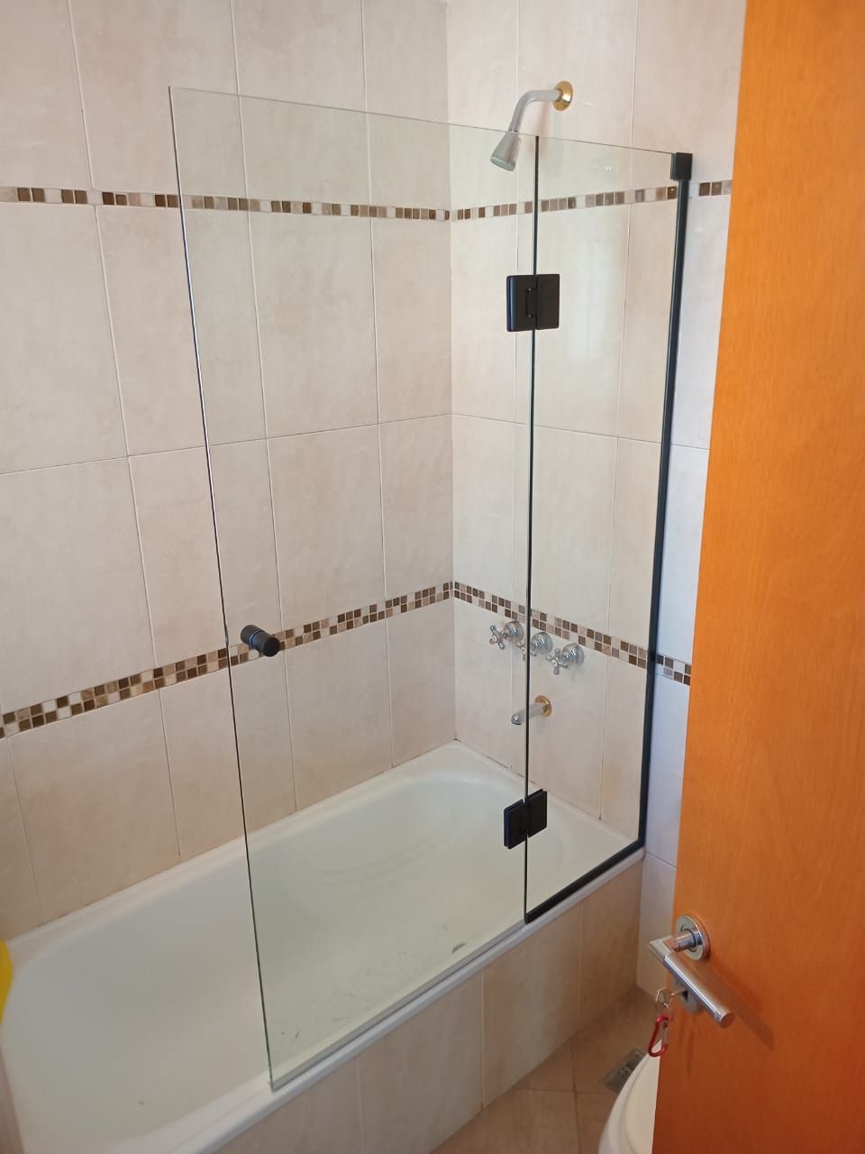 Ducha con puerta de cristal, adosada a bañera, con paredes de azulejos beige y puerta naranja.