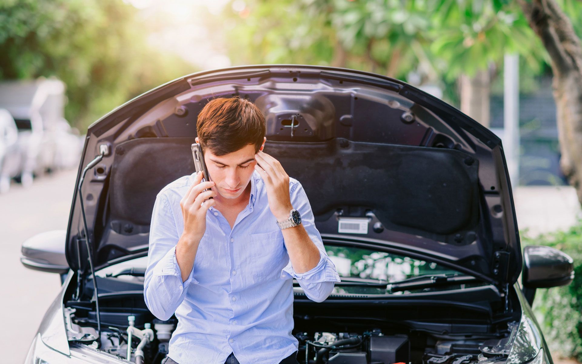 A man is sitting under the hood of a broken down car talking on a cell phone.
