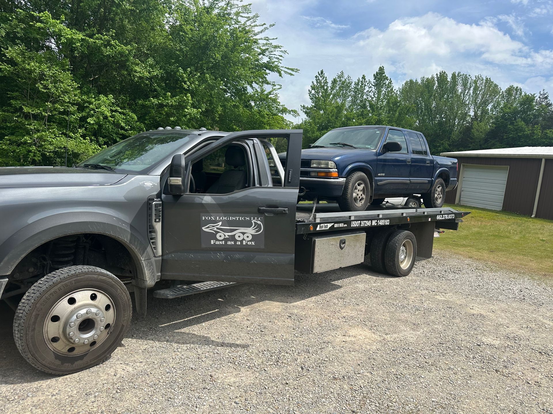 A tow truck with a blue truck on the back is parked in a gravel lot.