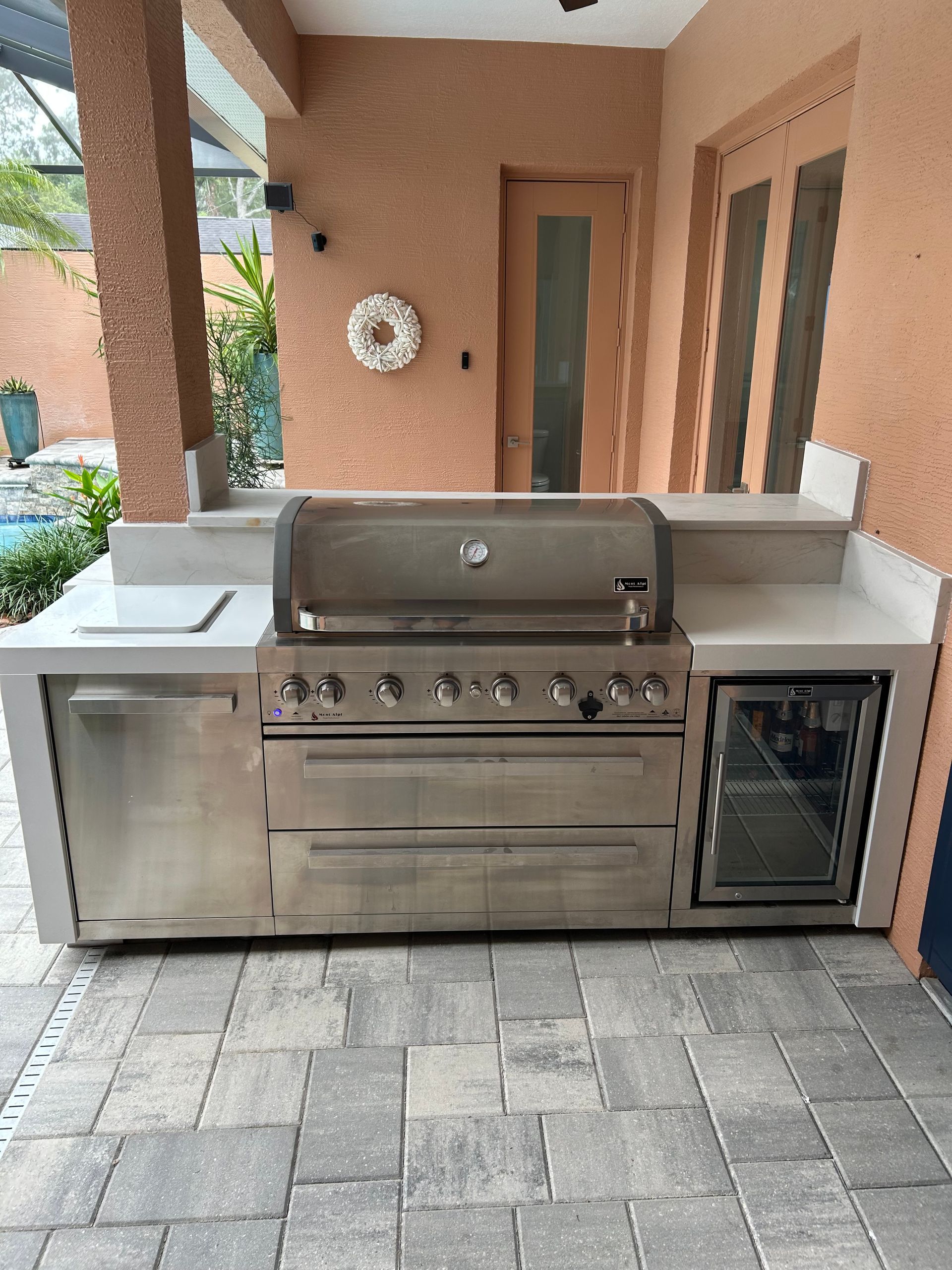 Outdoor kitchen with stainless steel grill, refrigerator, and cabinets on a tiled patio.