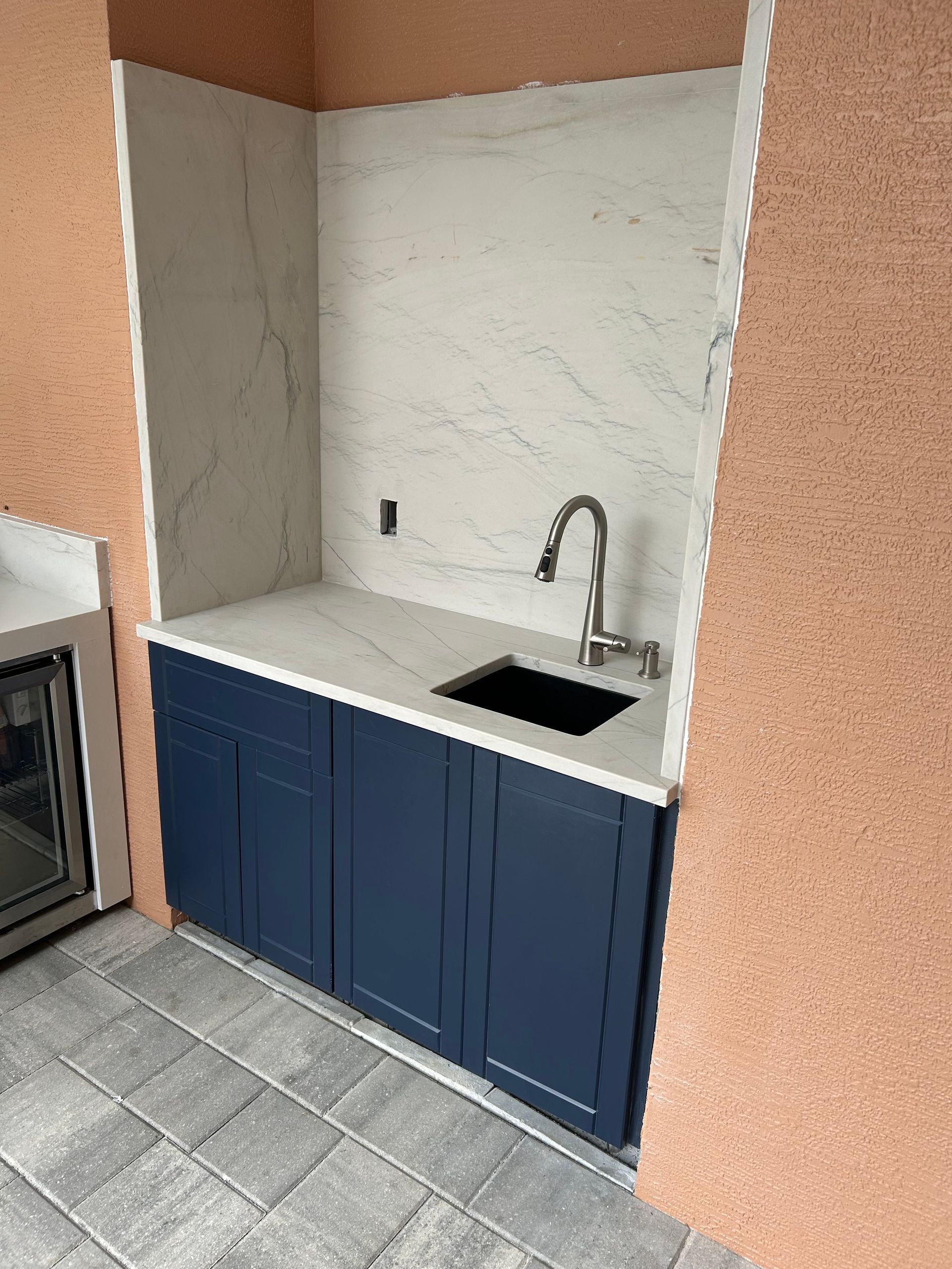 Built-in outdoor kitchen sink. Blue cabinets, white countertop, marble backsplash, and stainless steel faucet.
